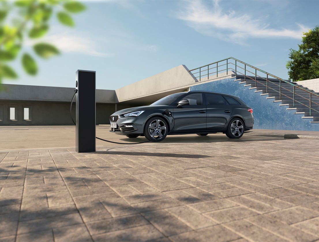 A grey SEAT Leon SP OW parked at an electric vehicle charging station. The car features sleek, aerodynamic lines, LED headlights and alloy wheels. The setting is modern, with a staircase and a clear blue sky in the background, highlighting the car's eco-friendly design and advanced technology.